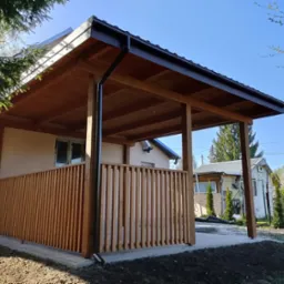 Drewniana altana z czarną rynną, osłonięta pionowymi listwami, przylegająca do budynku, z widocznym fragmentem ogrodu i niebem w tle.