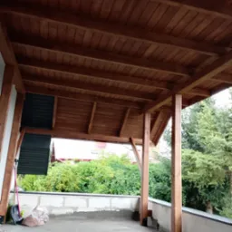 Zadaszony taras z drewnianą konstrukcją i widokiem na zieleń. Elementy konstrukcyjne w kolorze brązowym. Taras w trakcie wykańczania.