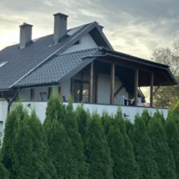 Dom z ciemną dachówką, widocznymi kominami i zadaszonym balkonem, otoczony zielonymi tujami. Ujęcie z dołu, w tle pochmurne niebo.