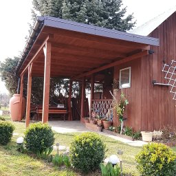 Usługi ogólnobudowlane - Drewniana pergola z czarnym dachem, przylegająca do budynku z dekoracjami. W tle zielone drzewo, a na pierwszym planie kuliste krzewy i lampki ogrodowe.