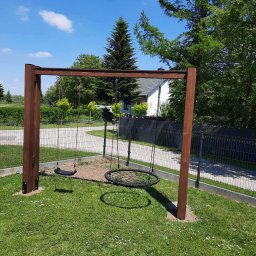 Usługi ogólnobudowlane - Drewniana konstrukcja huśtawek na trawniku, z widocznym siedziskiem typu bocianie gniazdo i pojedynczym siedziskiem. W tle dom i zieleń, błękitne niebo z chmurami.
