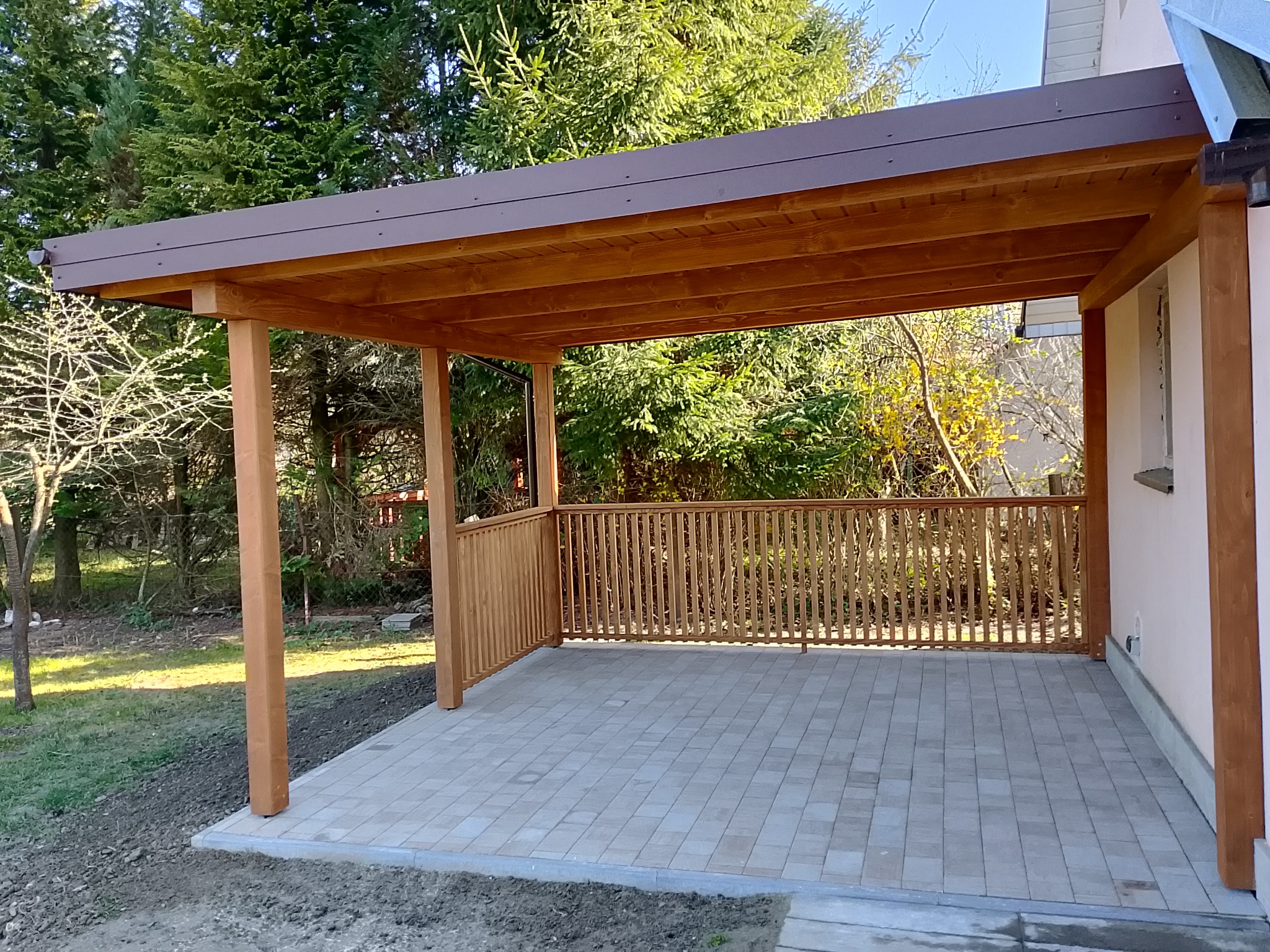 Zadaszony taras z drewnianą konstrukcją i brązowym dachem, wyłożony szarą kostką brukową, z widokiem na zielony ogród.