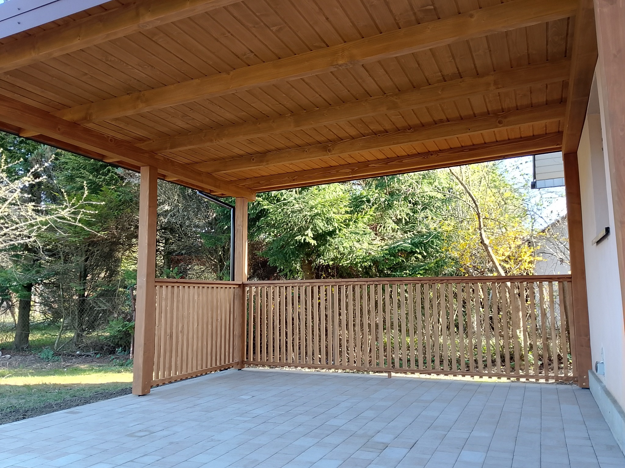 Zadaszony taras z drewnianą konstrukcją i balustradą, widok na ogród z kostką brukową na podłodze.