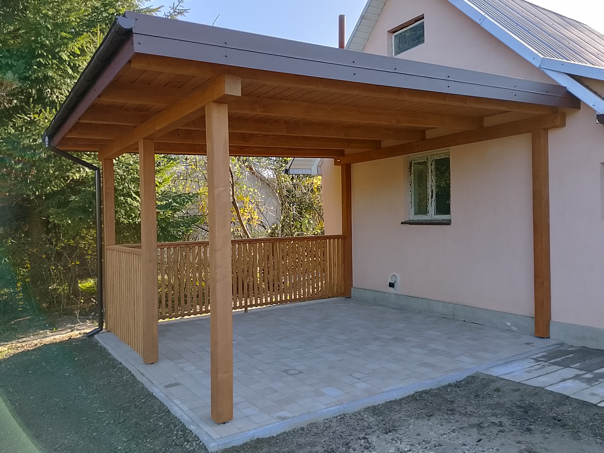 Drewniana wiata przylegająca do domu z szarą dachówką, wykończona ozdobną balustradą i wyłożona kostką brukową, widok z zewnątrz w słoneczny dzień.