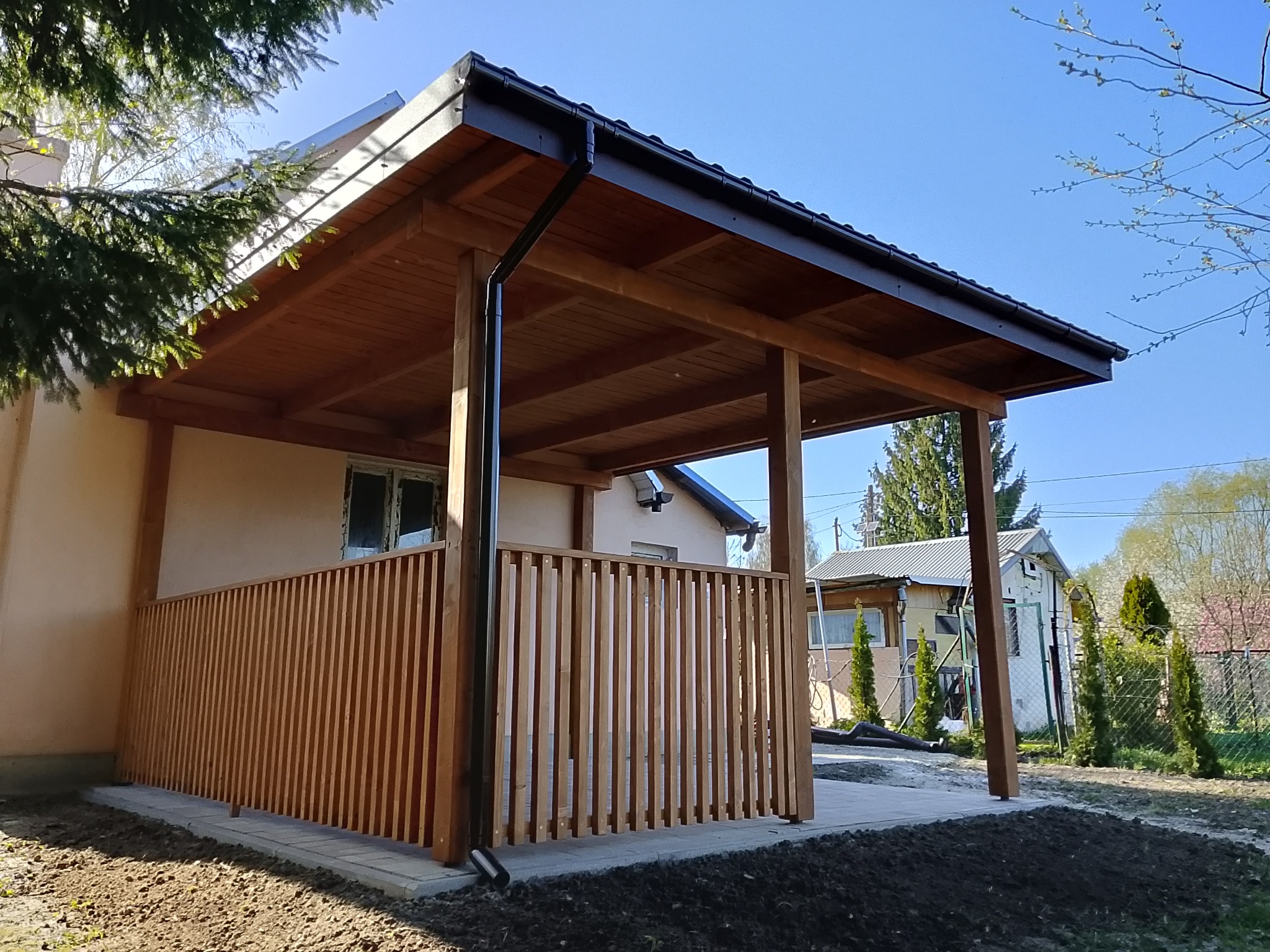Drewniana altana z czarną rynną, osłonięta pionowymi listwami, przylegająca do budynku, z widocznym fragmentem ogrodu i niebem w tle.