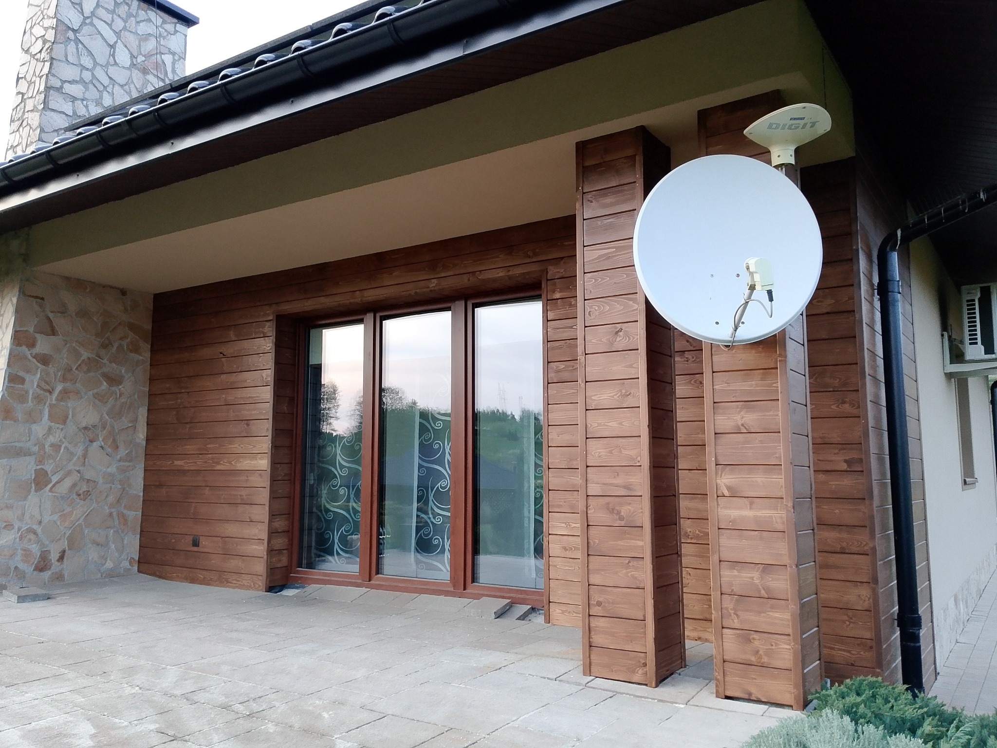 Drewniana elewacja domu z kamiennym kominem i tarasem. Widoczne brązowe deski, okna z dekoracyjnymi zasłonami, płytki tarasowe i antena satelitarna na elewacji.
