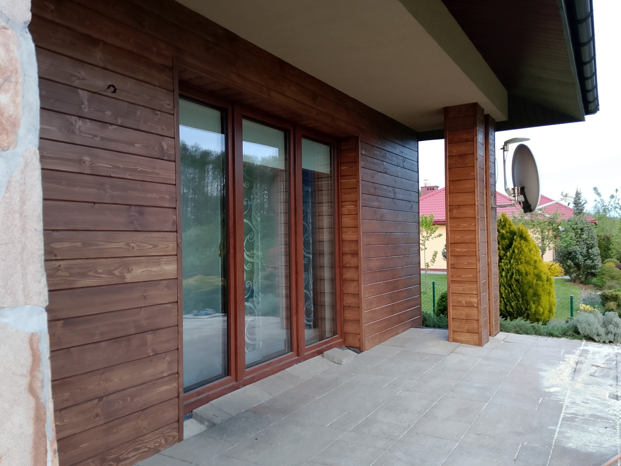 Drewniana elewacja domu z oknami i zadaszonym tarasem wyłożonym kostką. Widoczna antena satelitarna i zadbany ogród w tle.