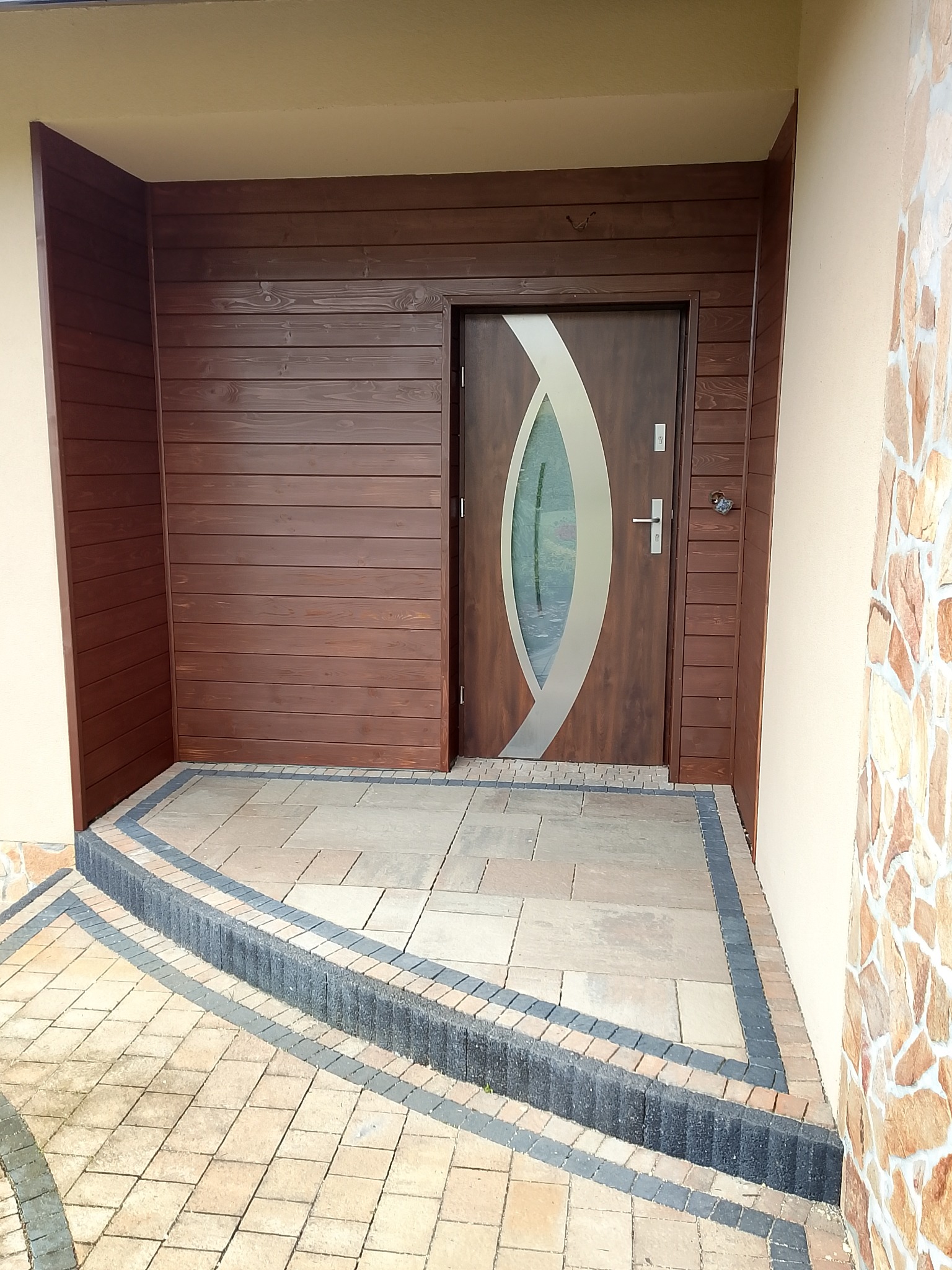 Wejście do domu z drewnianą boazerią i nowoczesnymi drzwiami z owalnym przeszkleniem. Uwagę przykuwa wykończenie schodów kostką brukową w dwóch odcieniach.