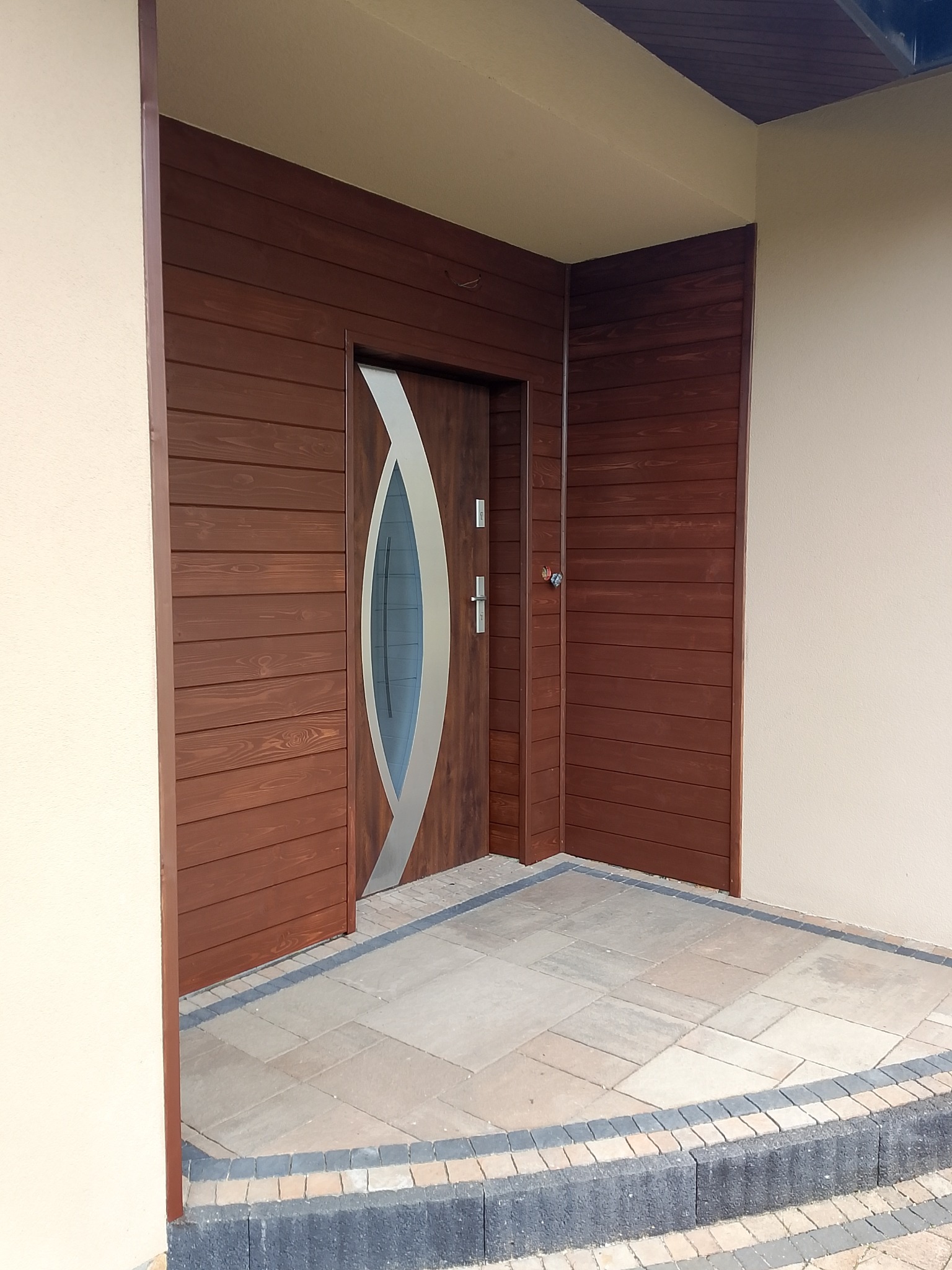 Nowoczesne drzwi wejściowe z drewnianą okładziną i elementami ze stali nierdzewnej, osadzone w elewacji wykończonej deską w ciepłym, brązowym odcieniu.