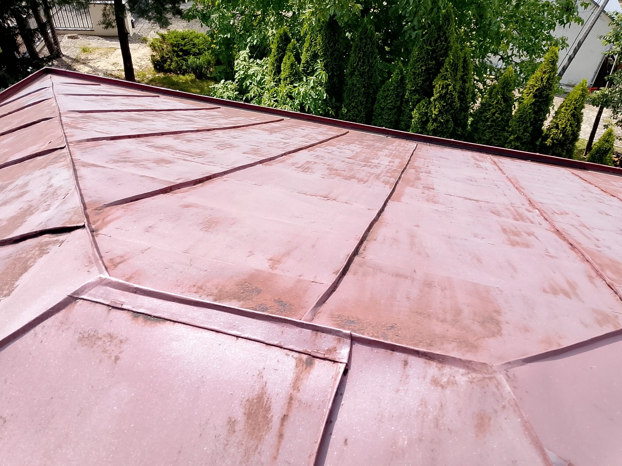 Metalowy dach w odcieniach czerwieni, widoczny z bliska, z zielenią drzew w tle. Ukazane detale łączeń blacharskich i faktura powierzchni.