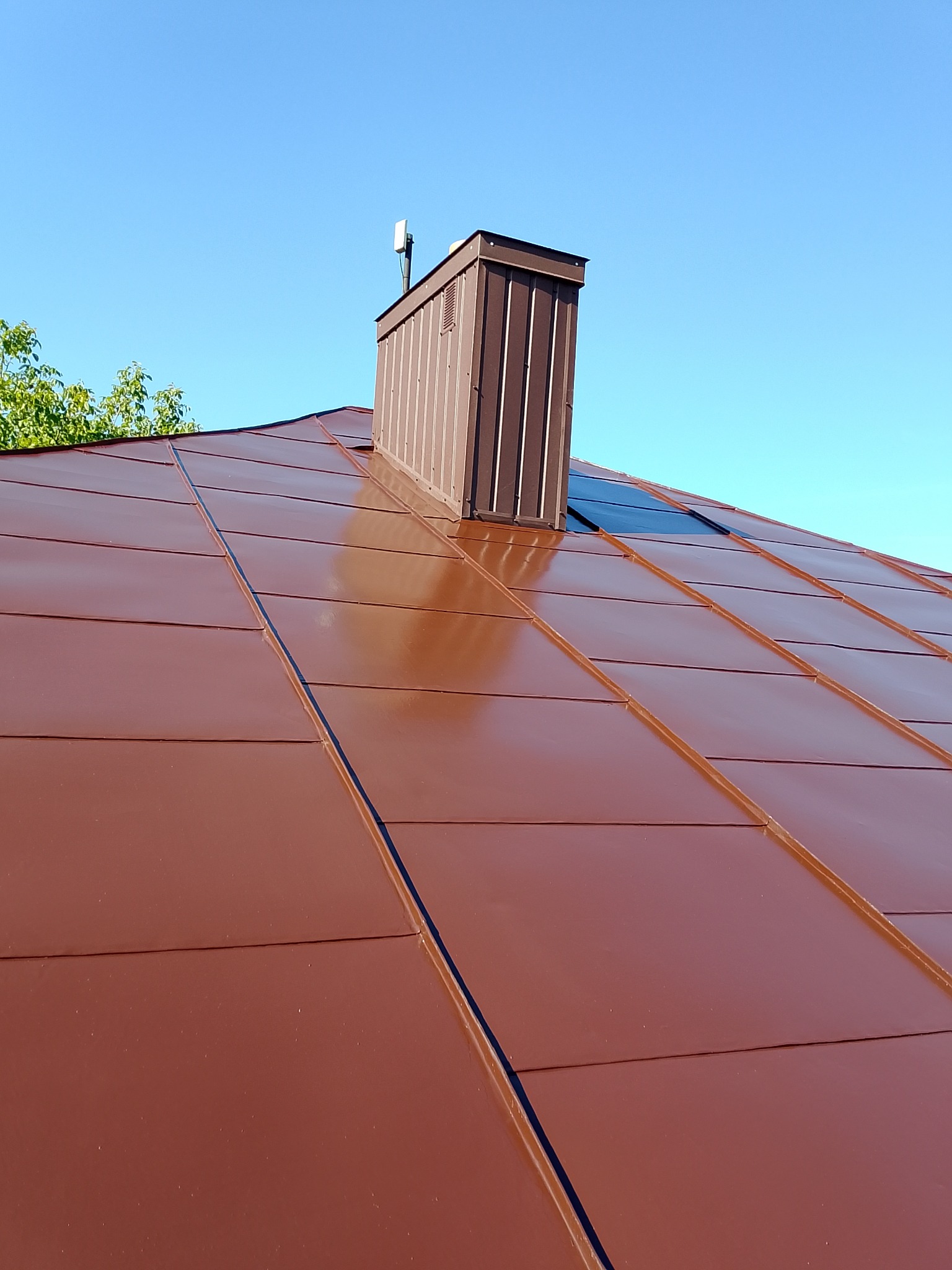 Błyszczący, brązowy dach z blachy trapezowej i kominem w kolorze miedzi na tle błękitnego nieba. Widoczny fragment zieleni. Nowe pokrycie dachowe.