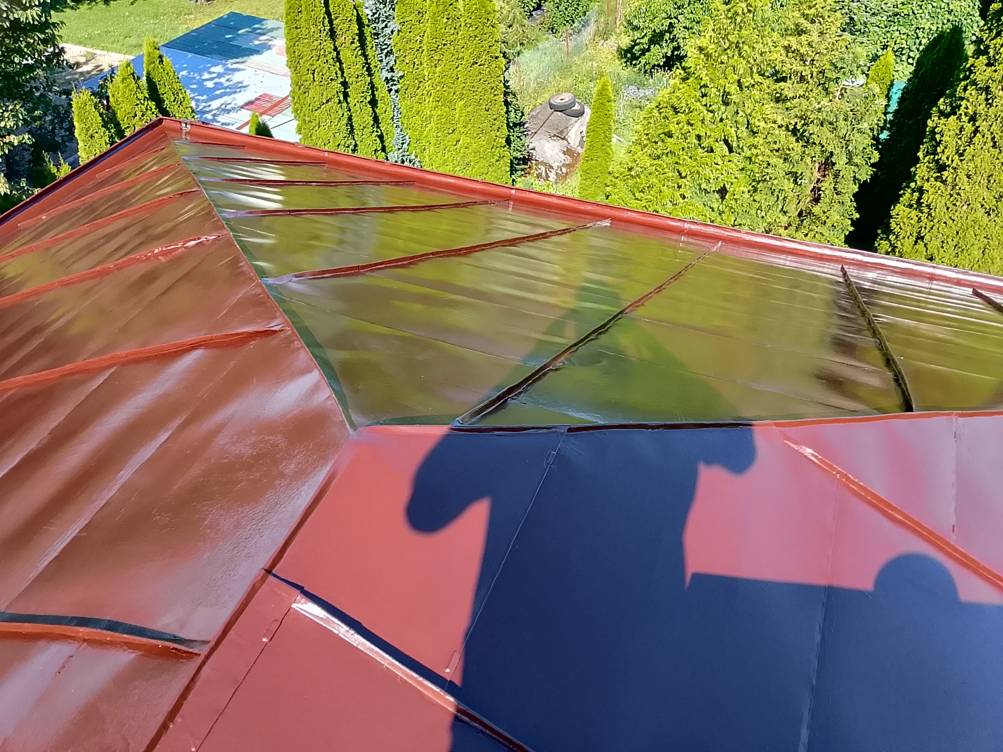 Czerwono-brązowy dach z blachy trapezowej, częściowo pokryty przezroczystą folią ochronną, z zielonym tłem drzew i krzewów. Widoczny cień osoby na dachu.