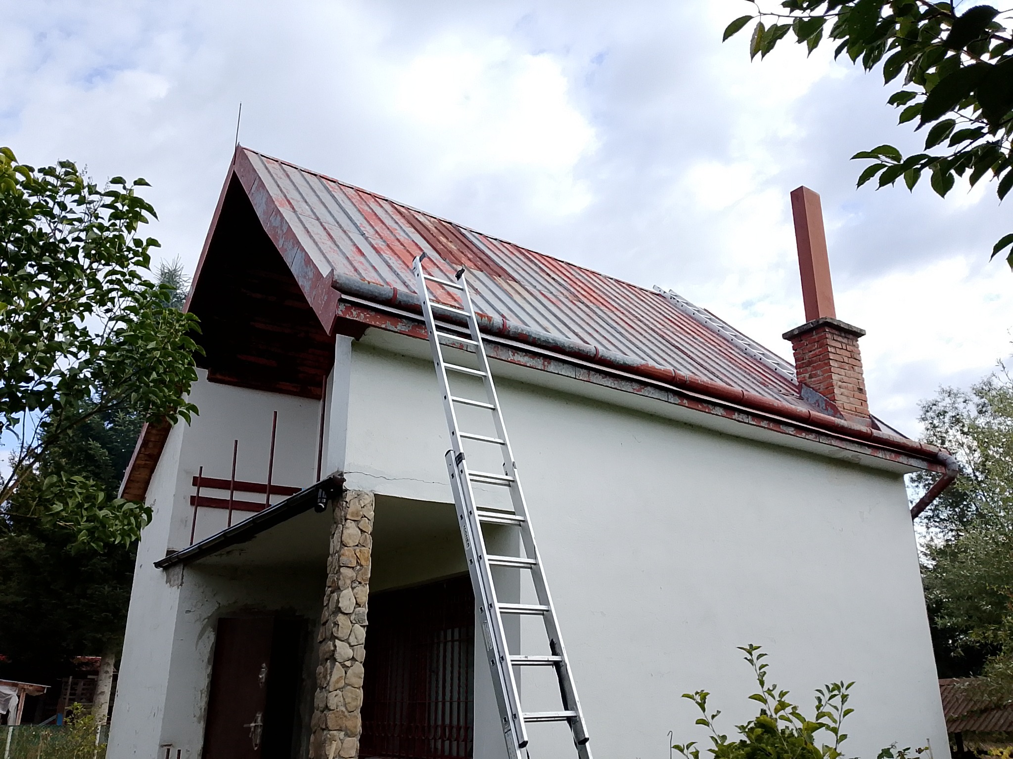 Dwukondygnacyjny dom z blachy falistej i kominem, z przystawioną aluminiową drabiną. Widoczne ślady korozji na dachu, częściowo odnowiona czerwień.