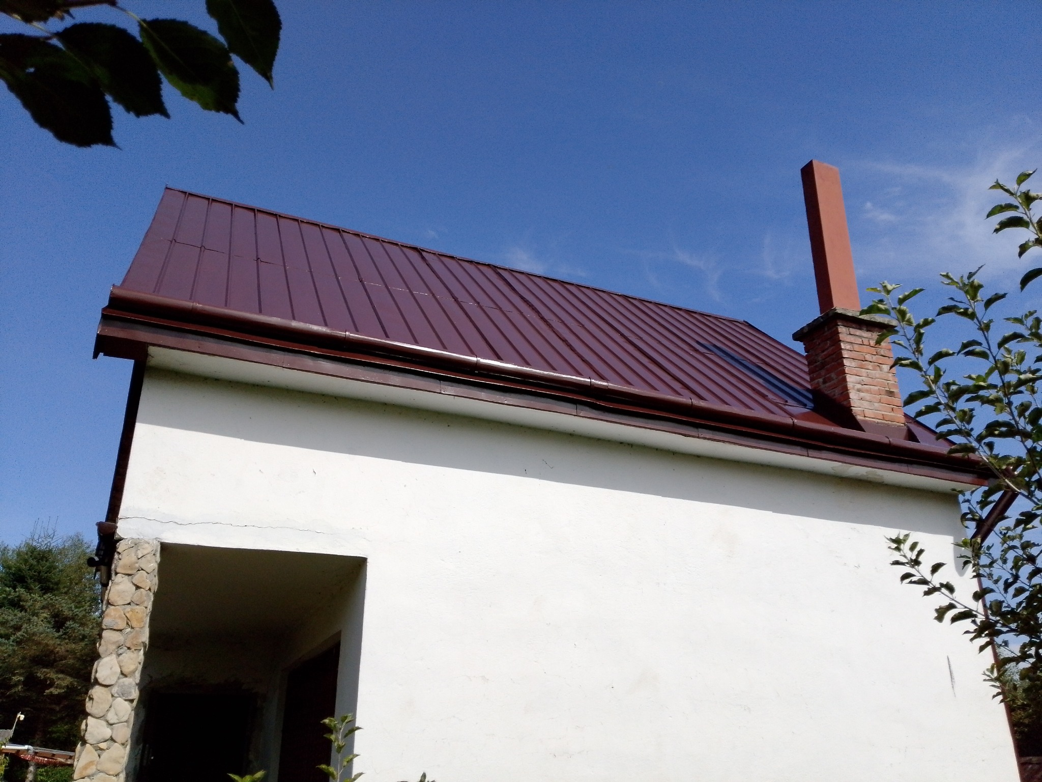 Dom z nowym, brązowym dachem z blachy trapezowej i ceglanym kominem na tle błękitnego nieba. Widoczne fragmenty zieleni.