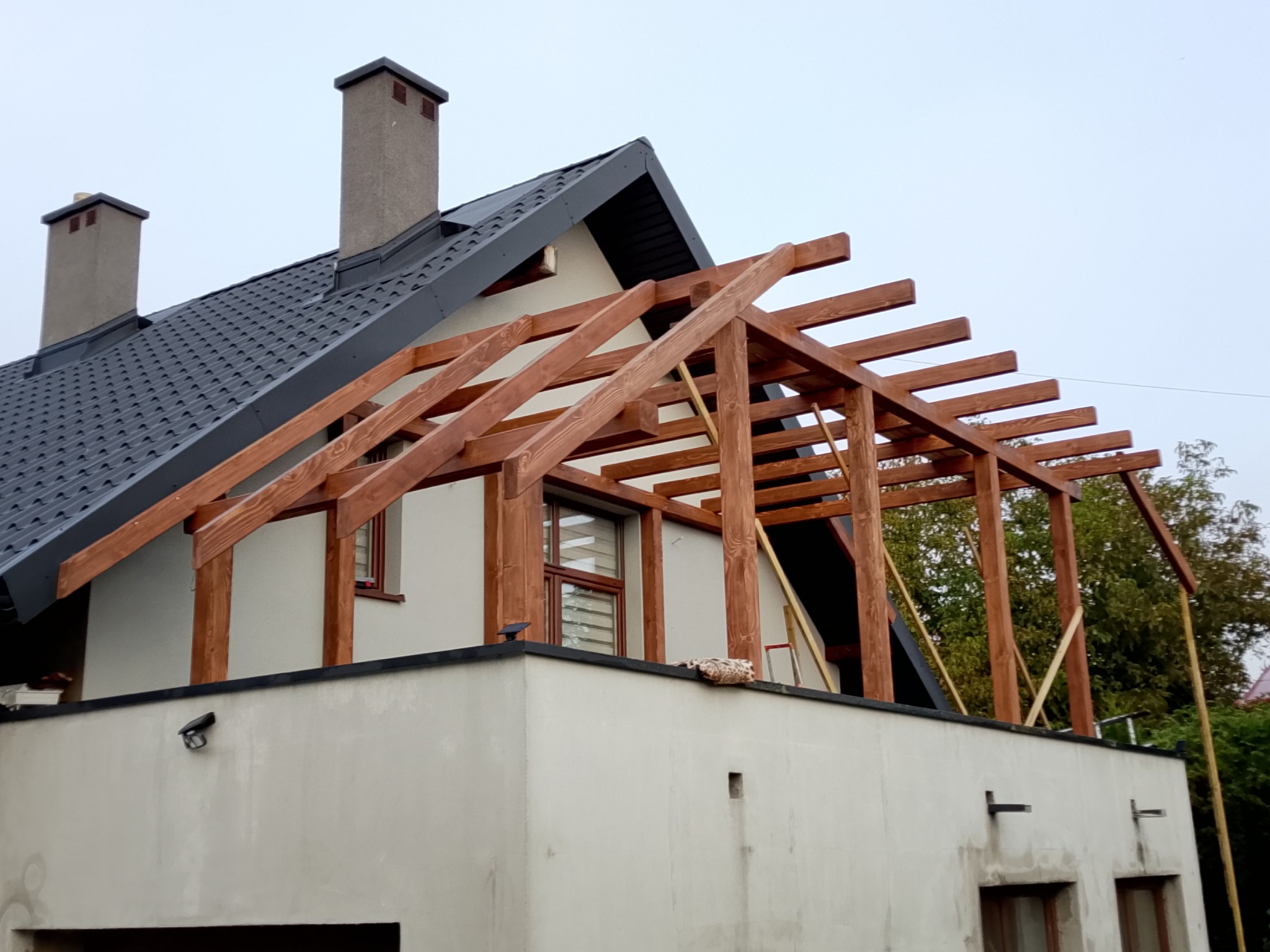 Konstrukcja drewnianej pergoli w trakcie montażu na balkonie domu z szarym dachem. Widoczne elementy mocujące i drabina. Pochmurne niebo w tle.