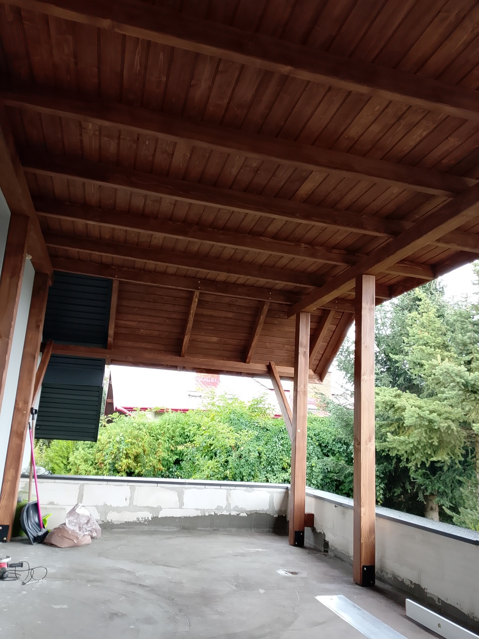 Zadaszony taras z drewnianą konstrukcją i widokiem na zieleń. Elementy konstrukcyjne w kolorze brązowym. Taras w trakcie wykańczania.