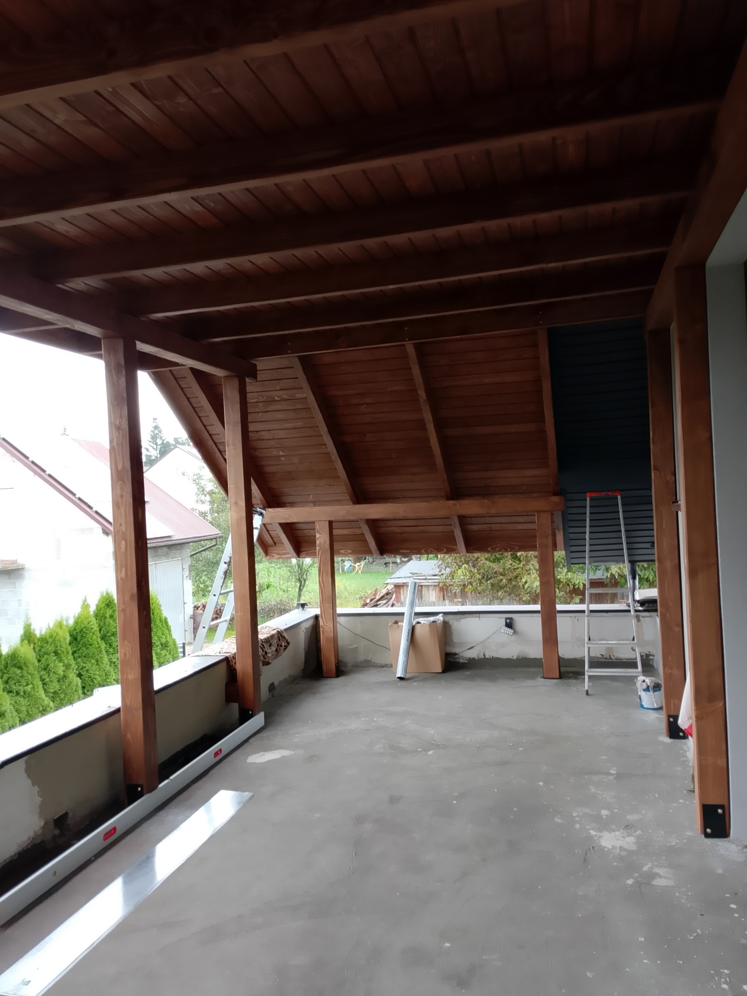 Zadaszony taras z drewnianą konstrukcją, widoczne słupy i deski. Na podłodze ślady prac remontowych, w tle drabina i elementy otoczenia.