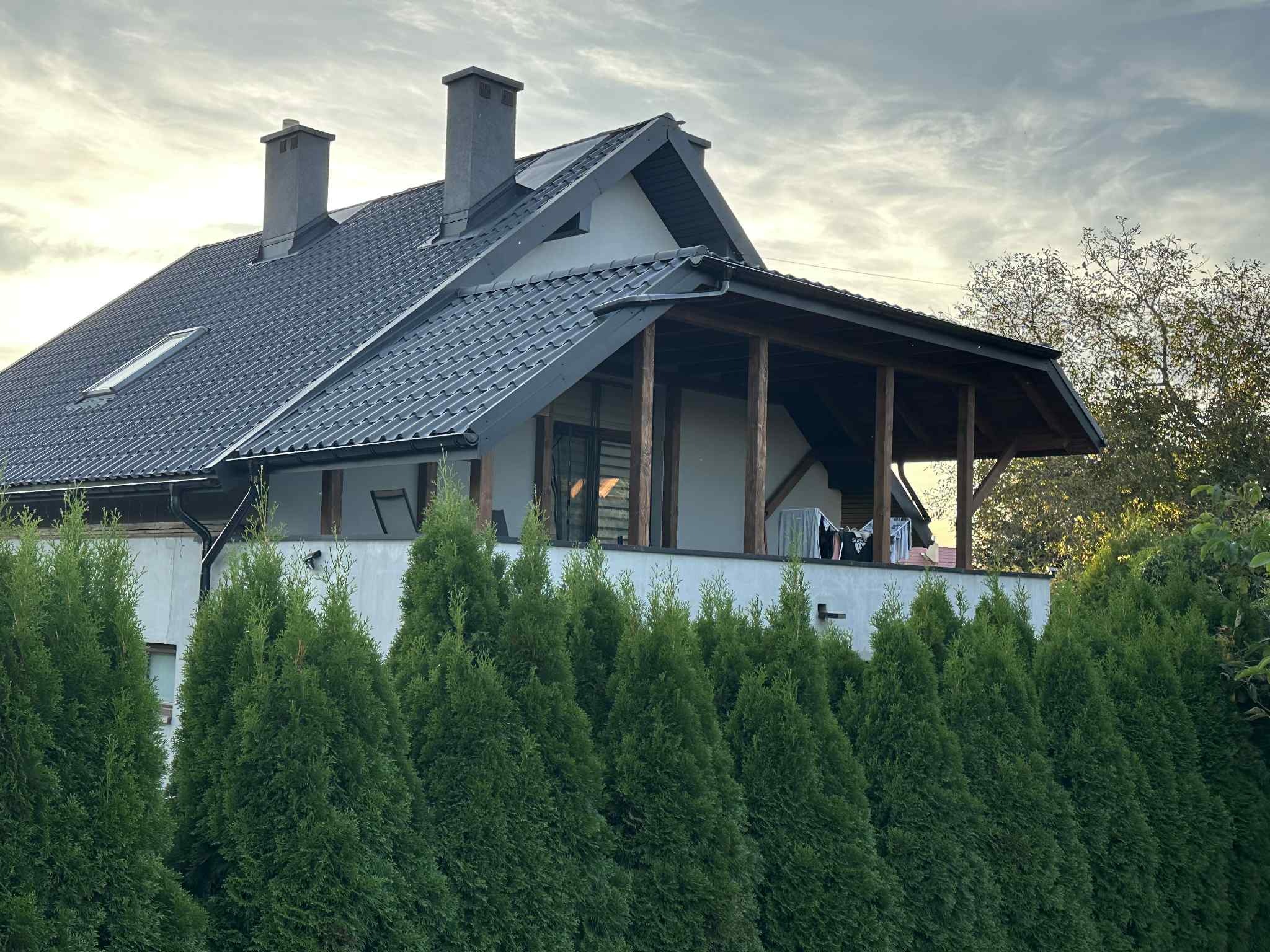 Dom z ciemną dachówką, widocznymi kominami i zadaszonym balkonem, otoczony zielonymi tujami. Ujęcie z dołu, w tle pochmurne niebo.