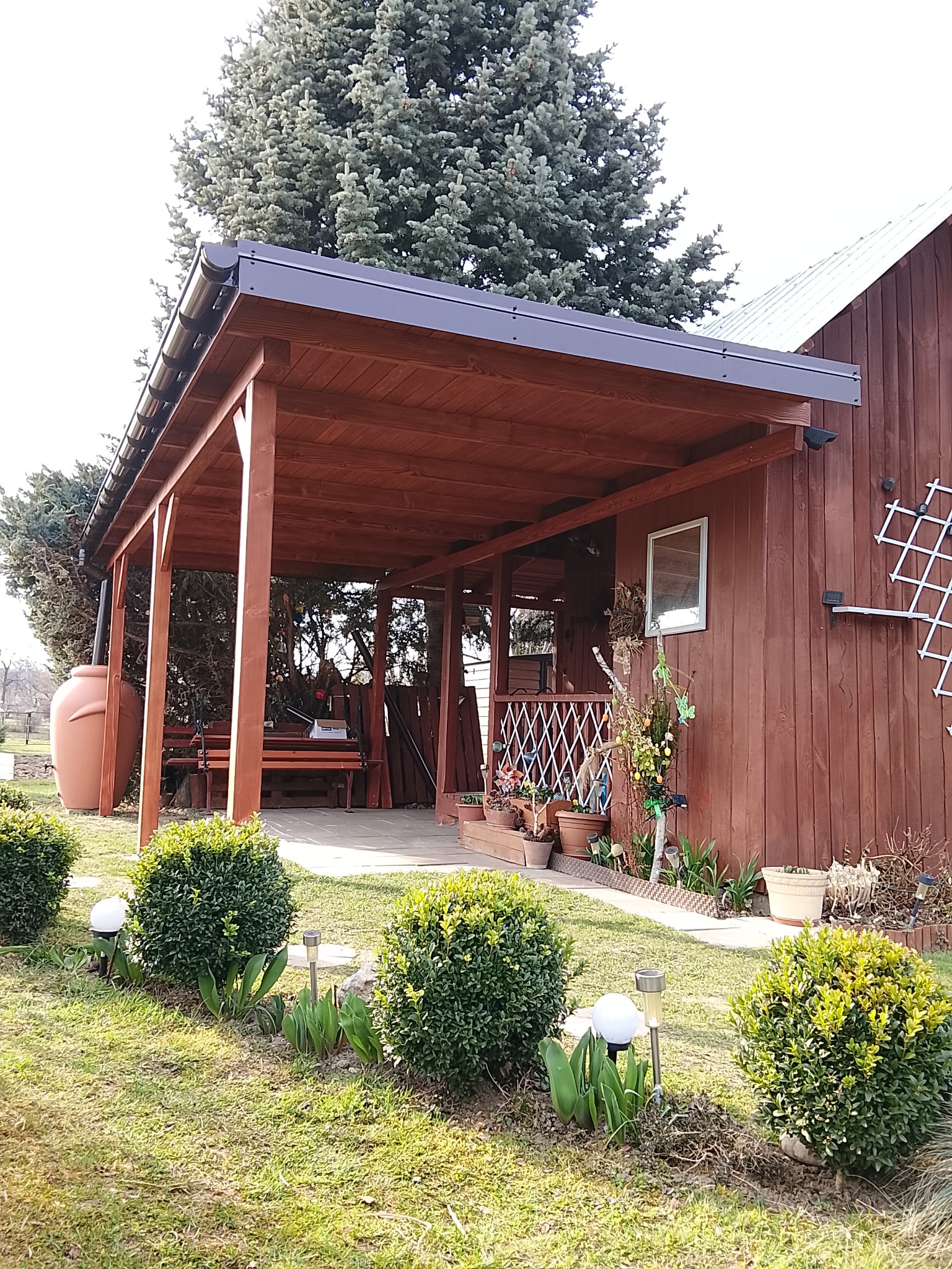 Drewniana pergola z czarnym dachem, przylegająca do budynku z dekoracjami. W tle zielone drzewo, a na pierwszym planie kuliste krzewy i lampki ogrodowe.