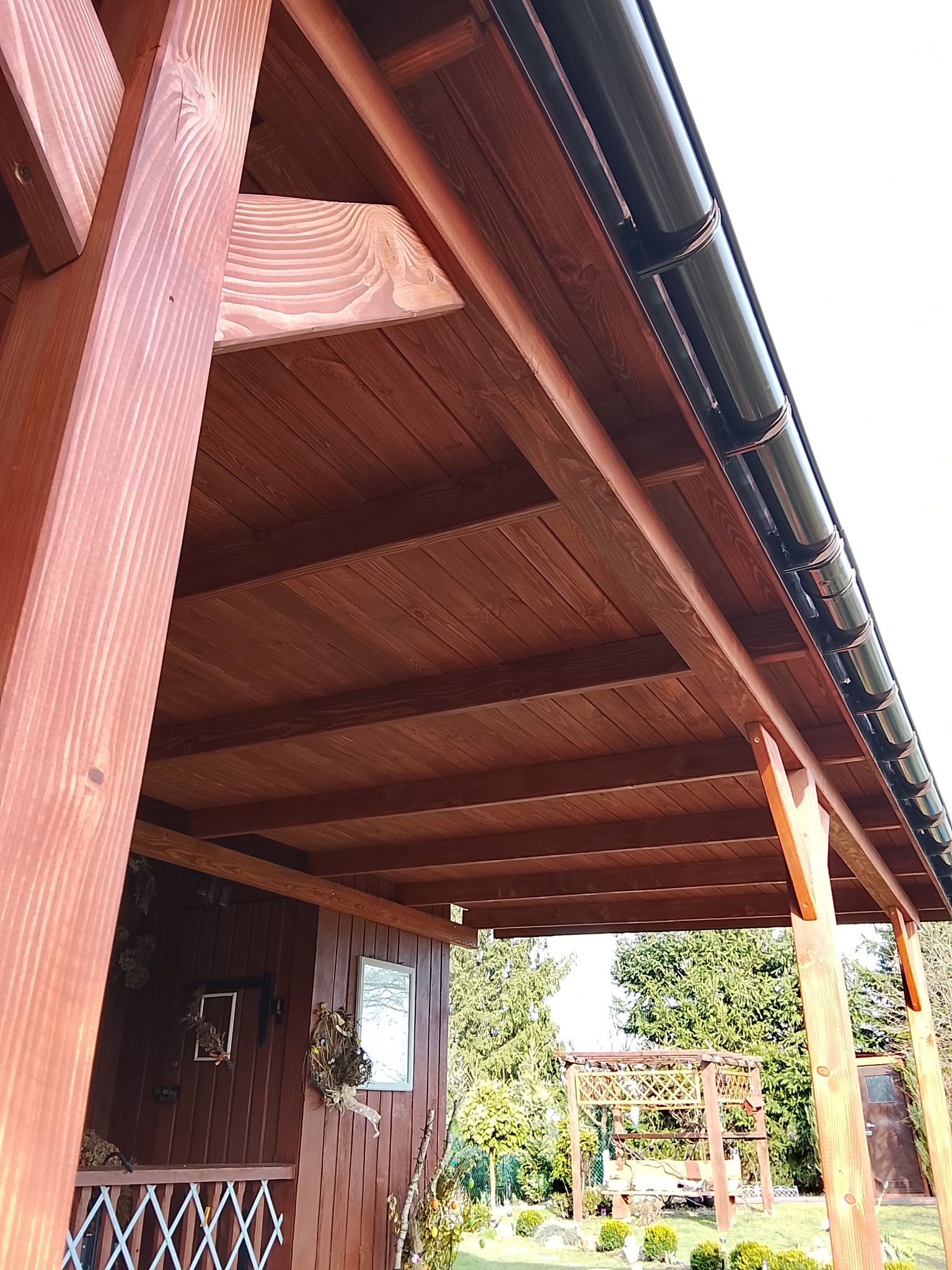 Drewniana pergola z ciemnobrązowym dachem i czarnymi rynnami, widok z dołu, ukazujący konstrukcję i detale wykończenia. W tle zieleń ogrodu i elementy dekoracyjne.