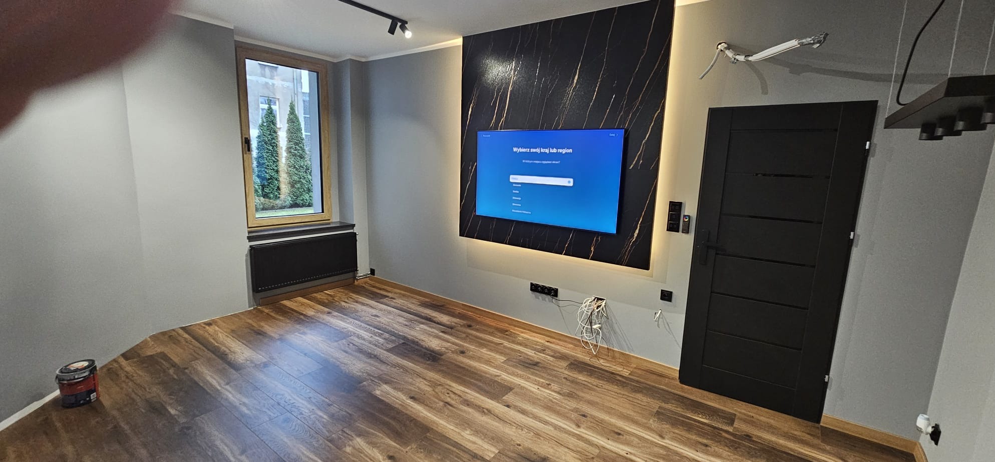 Pokój po remoncie: drewnopodobna podłoga, szare ściany, okno z widokiem na zieleń, czarny panel z podświetleniem LED za telewizorem, czarne drzwi.
