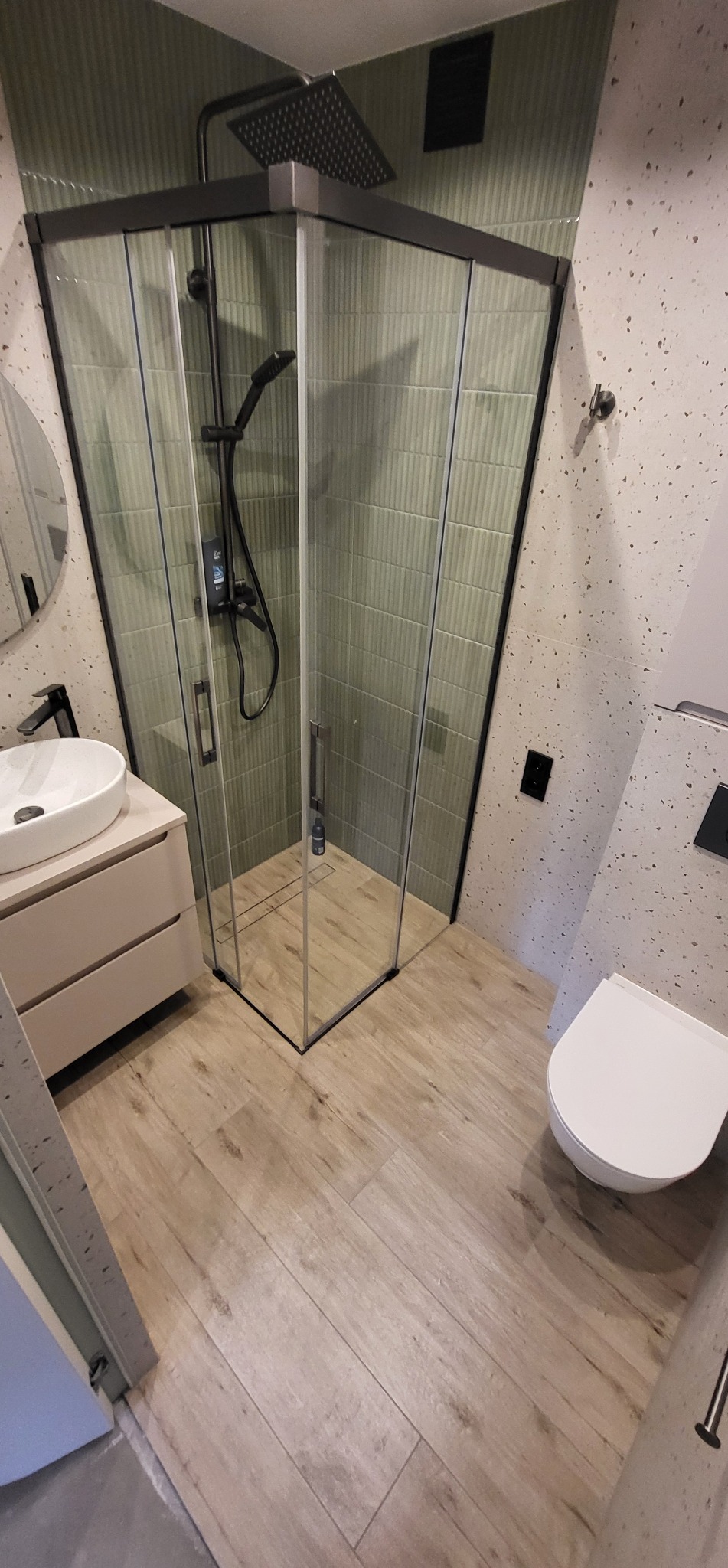 Nowoczesna łazienka z kabiną prysznicową typu walk-in, umywalką na szafce i toaletą. Ściany wykończone płytkami w odcieniach zieleni i beżu. Drewnopodobna podłoga.