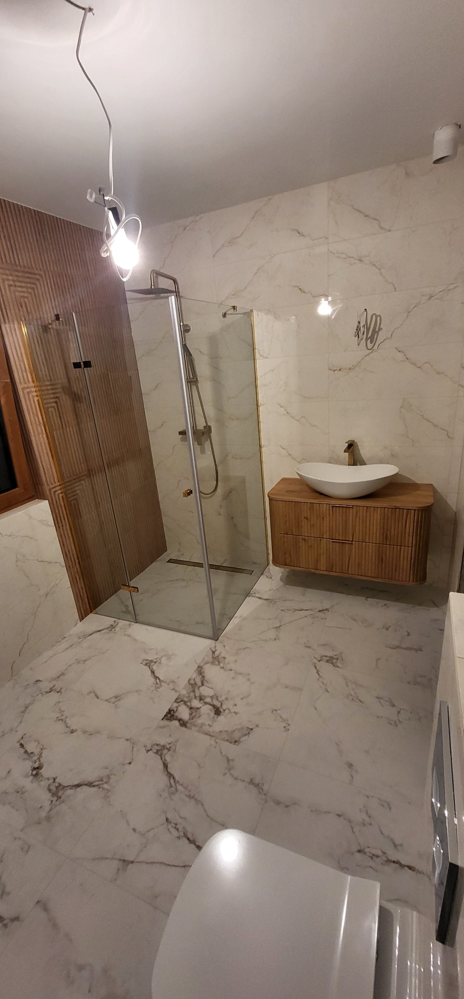 Nowoczesna łazienka z kabiną prysznicową typu walk-in, umywalką nablatową na drewnianej szafce i marmurową podłogą. Widoczne detale wykończenia.