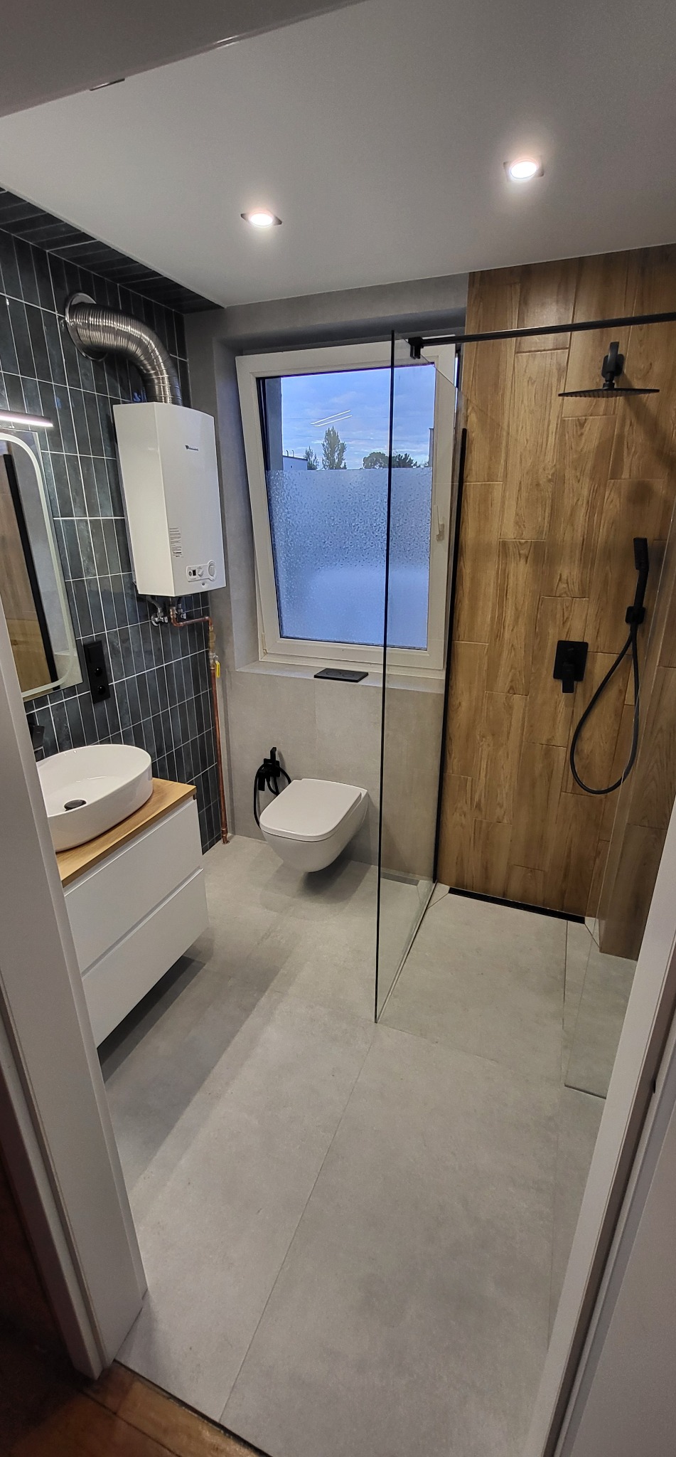 Nowoczesna łazienka z kabiną typu walk-in, umywalką nablatową i grzejnikiem. Ciemne płytki na ścianie kontrastują z drewnopodobnymi w strefie prysznica.