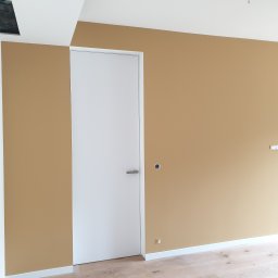 Kamil Ficner - Ściana pomalowana na kolor piaskowy z widoczną białą, minimalistyczną ościeżnicą i drzwiami oraz fragmentem drewnianej podłogi; widoczne niedokończone gniazdka elektryczne.