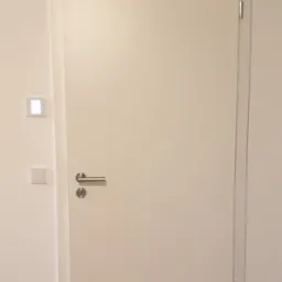 Minimalistyczne, białe drzwi wewnętrzne z ukrytymi zawiasami i srebrną klamką, zamontowane w ścianie z widocznym gniazdkiem elektrycznym i panelem sterowania oświetleniem.