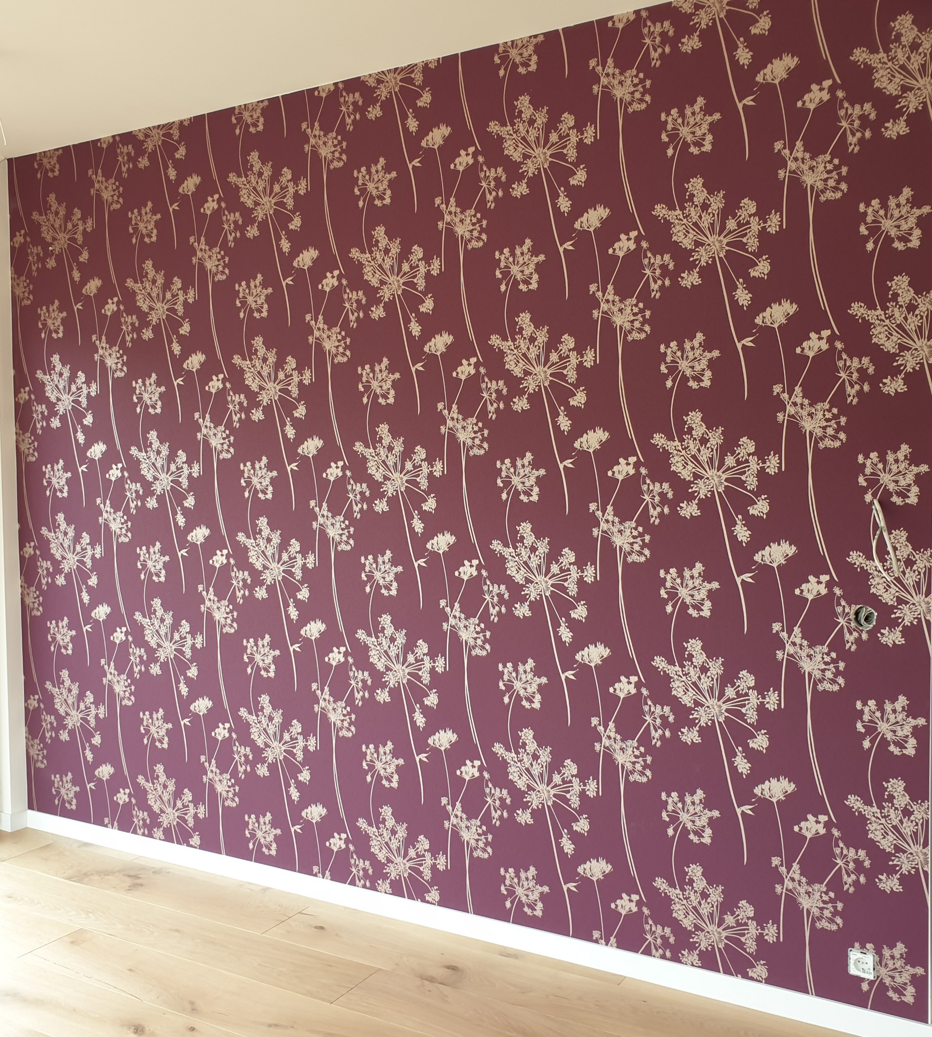 Ściana w pokoju pokryta bordową tapetą z delikatnym, kremowym wzorem roślinnym, widoczne jasne panele podłogowe i gniazdko elektryczne.