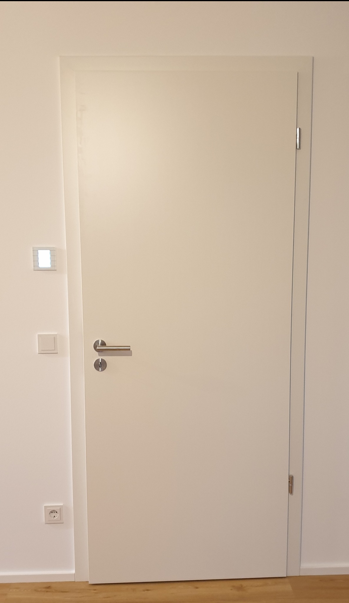 Minimalistyczne, białe drzwi wewnętrzne z ukrytymi zawiasami i srebrną klamką, zamontowane w ścianie z widocznym gniazdkiem elektrycznym i panelem sterowania oświetleniem.