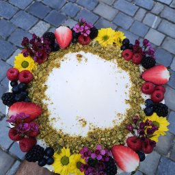 PRACOWNIA DOBROCI SPÓŁKA Z OGRANICZONĄ ODPOWIEDZIALNOŚCIĄ - Okrągły tort z białym kremem, pistacjową posypką i dekoracją z owoców (truskawki, maliny, jagody, jeżyny) oraz kwiatów na tle szarej kostki brukowej.