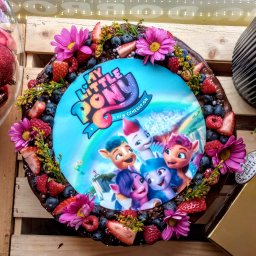 PRACOWNIA DOBROCI SPÓŁKA Z OGRANICZONĄ ODPOWIEDZIALNOŚCIĄ - Okrągły tort czekoladowy z motywem 'My Little Pony', udekorowany świeżymi owocami, kwiatami i srebrnymi perełkami, na drewnianym tle z dekoracjami świątecznymi.