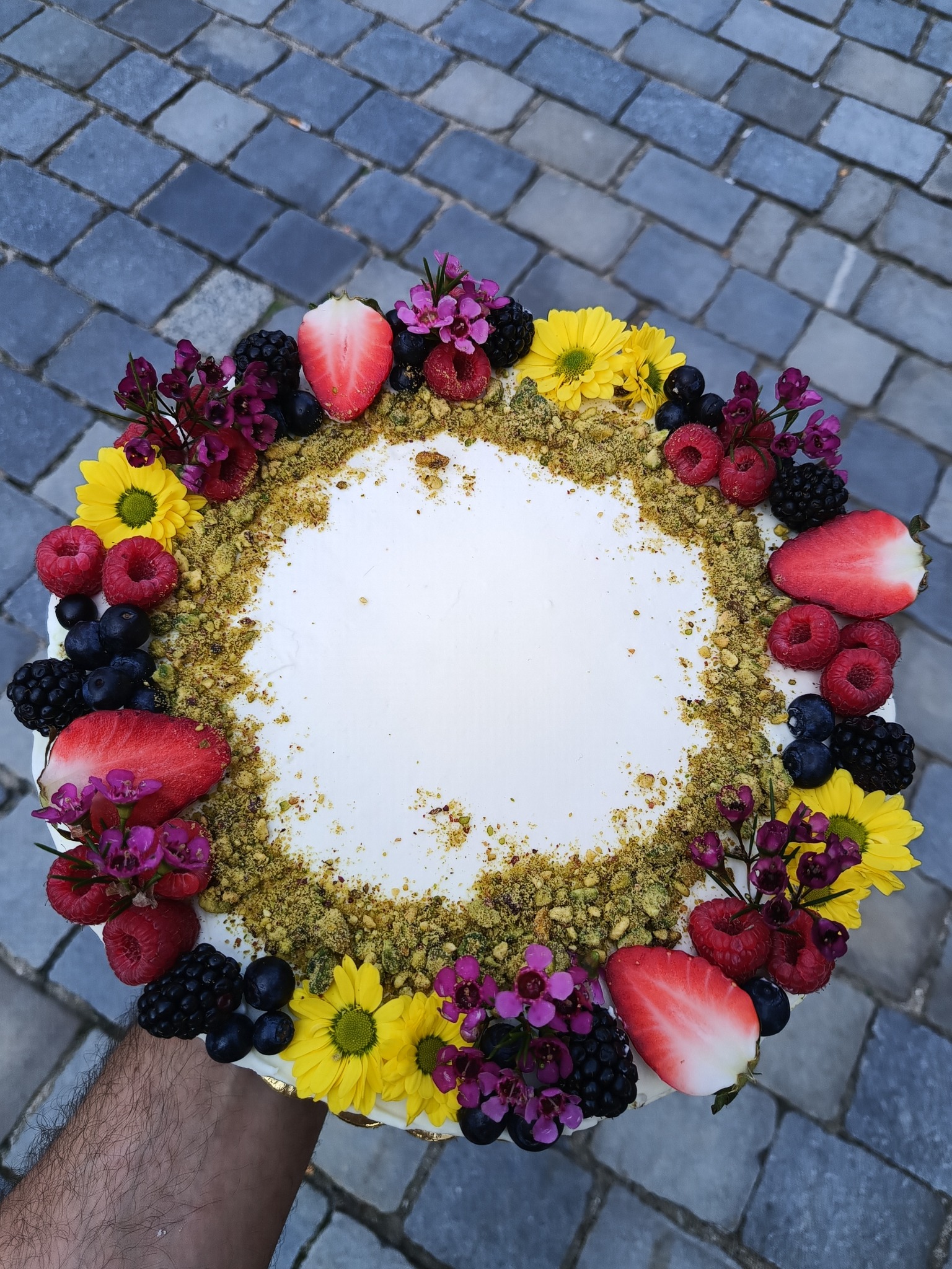 Okrągły tort z białym kremem, pistacjową posypką i dekoracją z owoców (truskawki, maliny, jagody, jeżyny) oraz kwiatów na tle szarej kostki brukowej.