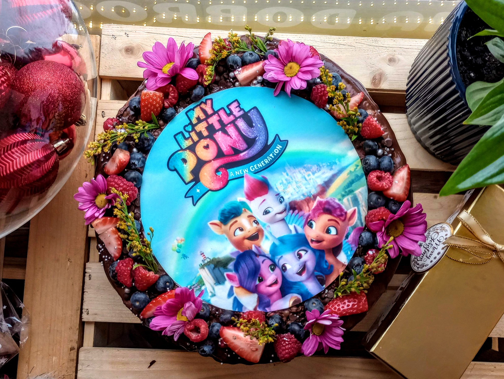 Okrągły tort czekoladowy z motywem 'My Little Pony', udekorowany świeżymi owocami, kwiatami i srebrnymi perełkami, na drewnianym tle z dekoracjami świątecznymi.