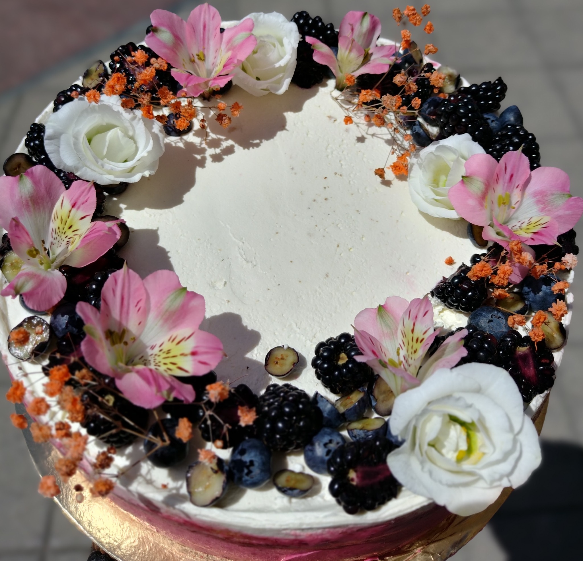 Kremowy tort z dekoracją z białych i różowych kwiatów, jeżyn, borówek i pomarańczowych gałązek. Widok z góry, delikatne światło podkreśla detale.