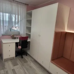 Pokój w odcieniach różu z białą szafą wnękową, biurkiem i tapicerowanym siedziskiem. Widoczne okno z firanką i dziecięce akcesoria.