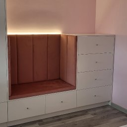 MARTINOS MEBLE NA WYMIAR Marcin Hełmiński - Biała zabudowa meblowa z siedziskiem wykończonym różowym welurem, oświetlona od góry. Szuflady z kryształowymi gałkami, parkiet na podłodze, różowe ściany.