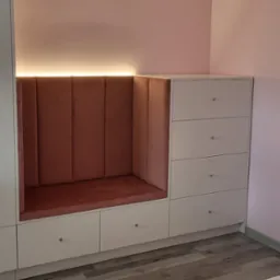 Biała zabudowa meblowa z siedziskiem wykończonym różowym welurem, oświetlona od góry. Szuflady z kryształowymi gałkami, parkiet na podłodze, różowe ściany.