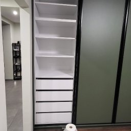 MARTINOS MEBLE NA WYMIAR Marcin Hełmiński - Nowoczesna szafa wnękowa z przesuwnymi drzwiami, białe półki i szuflady, czarne obramowanie. Na podłodze rolka papieru toaletowego i talerz po posiłku.