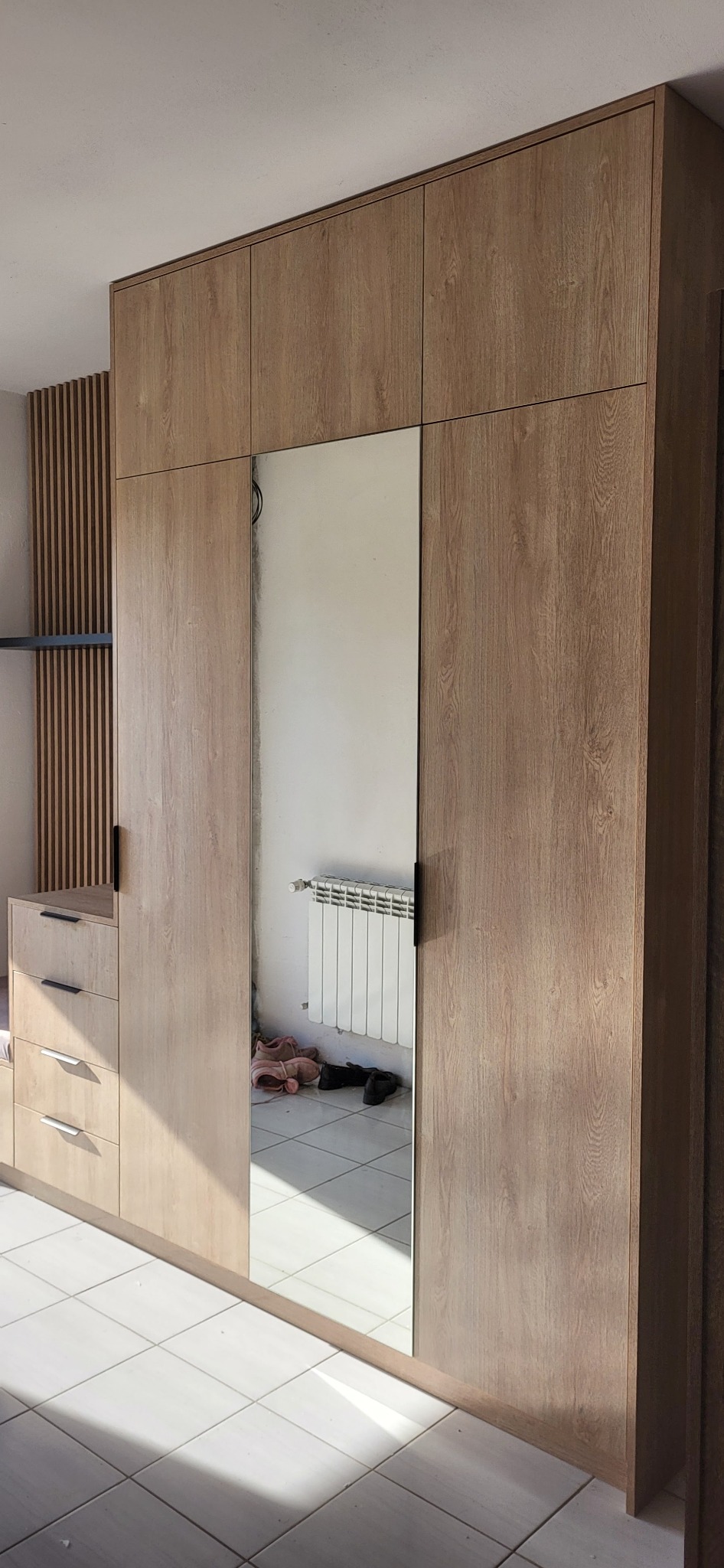 Drewniana szafa z lustrem, widoczne odbicie grzejnika i podłogi. Szafa z szufladami i pionowymi panelami dekoracyjnymi. Jasne płytki podłogowe.