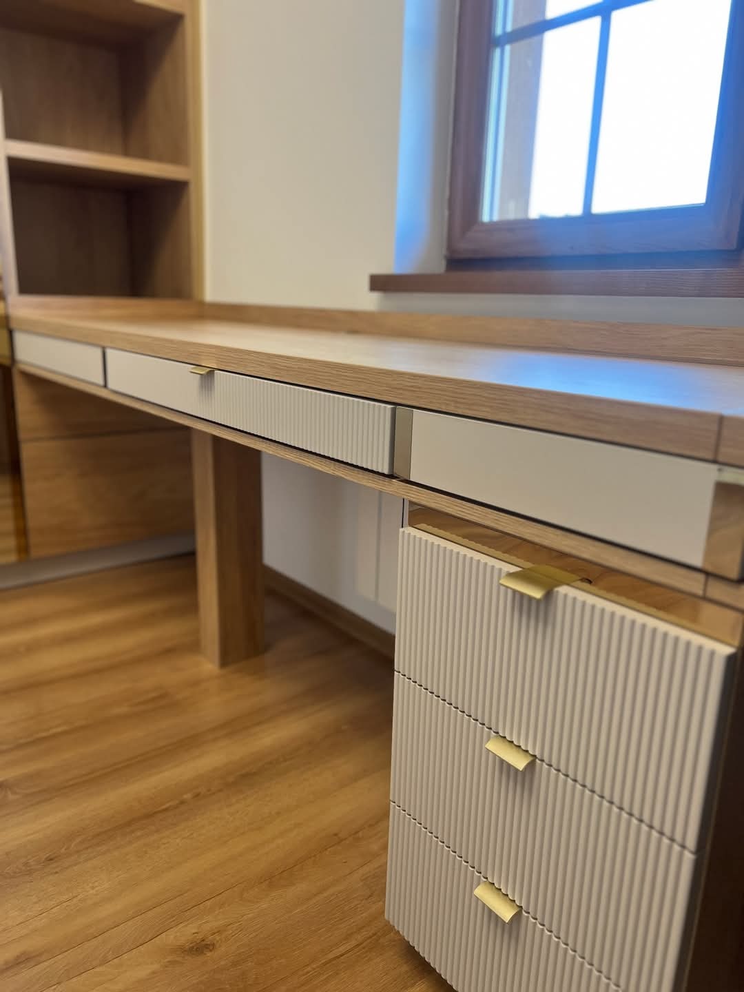 Biurko z drewna i białych frezowanych frontów szuflad, z złotymi uchwytami, wbudowane w regał, oświetlone światłem dziennym z okna w tle.