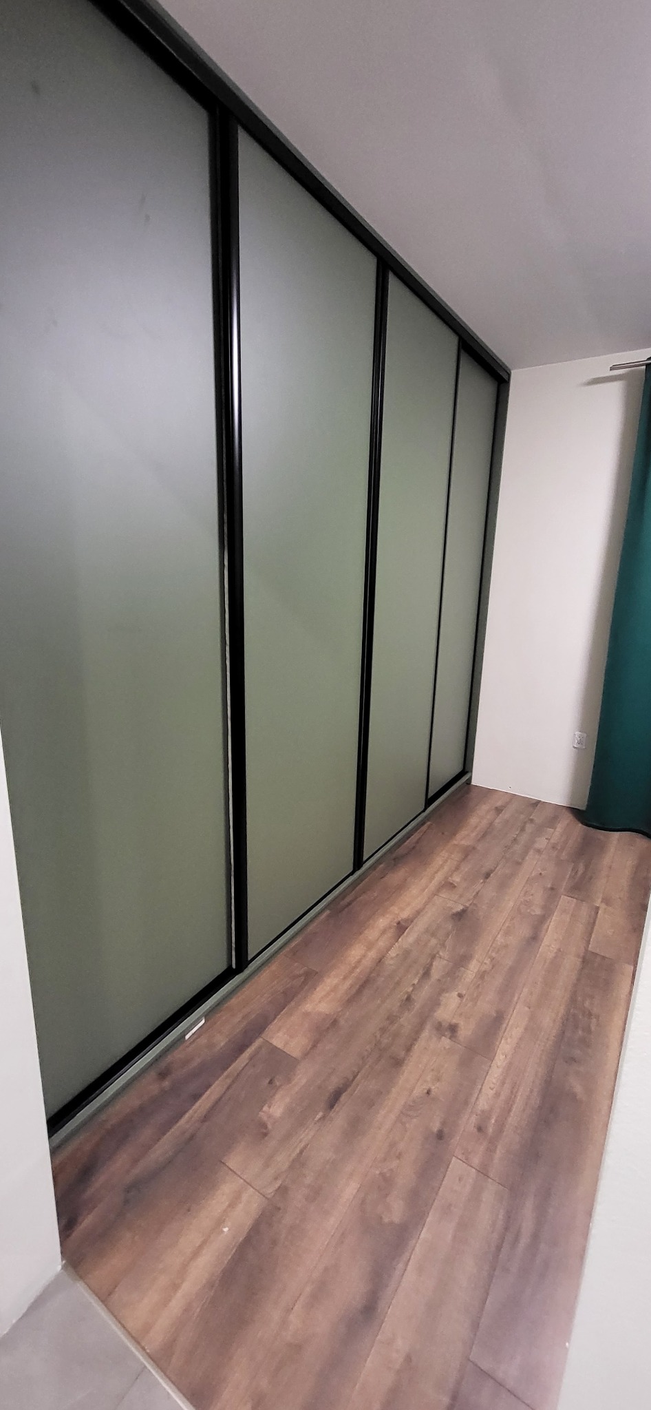 Szafa wnękowa z przesuwnymi, matowymi drzwiami i czarnymi ramami, widoczna na tle drewnianej podłogi i białej ściany z zieloną zasłoną.