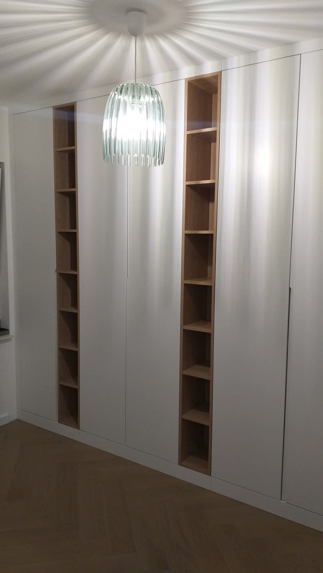 Biała szafa wnękowa z drewnianymi półkami, oświetlona lampą z dekoracyjnym kloszem, na parkiecie ułożonym w jodełkę. Minimalistyczny design.