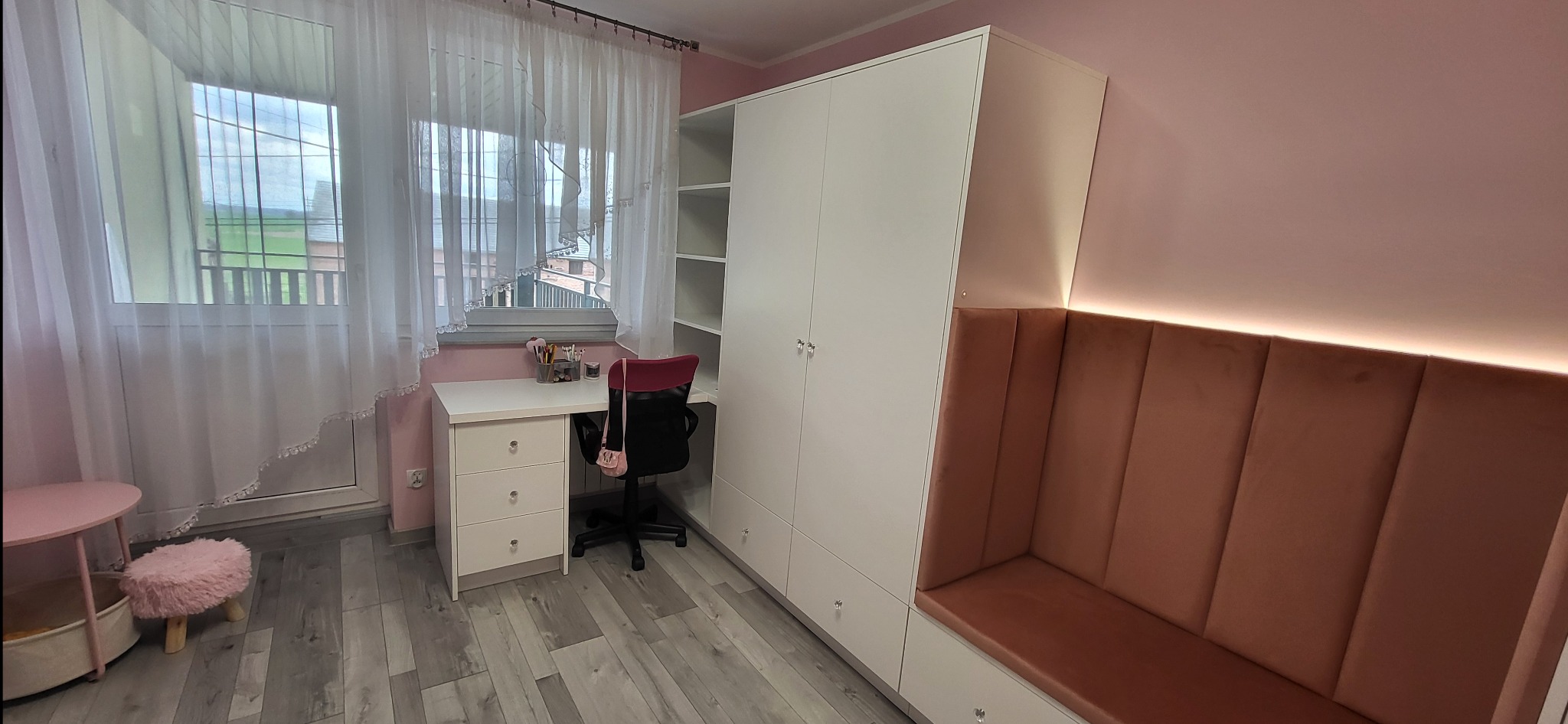 Pokój w odcieniach różu z białą szafą wnękową, biurkiem i tapicerowanym siedziskiem. Widoczne okno z firanką i dziecięce akcesoria.