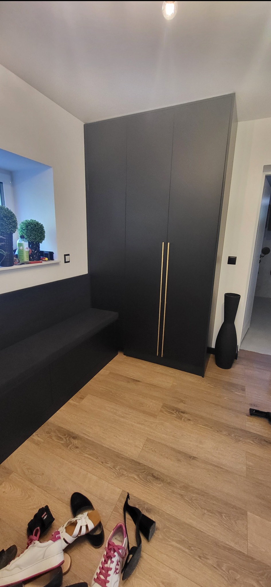 Czarna szafa wnękowa z złotymi uchwytami i siedziskiem w pokoju. Na podłodze leżą buty. Minimalistyczny design, jasne wnętrze, doniczki na parapecie.