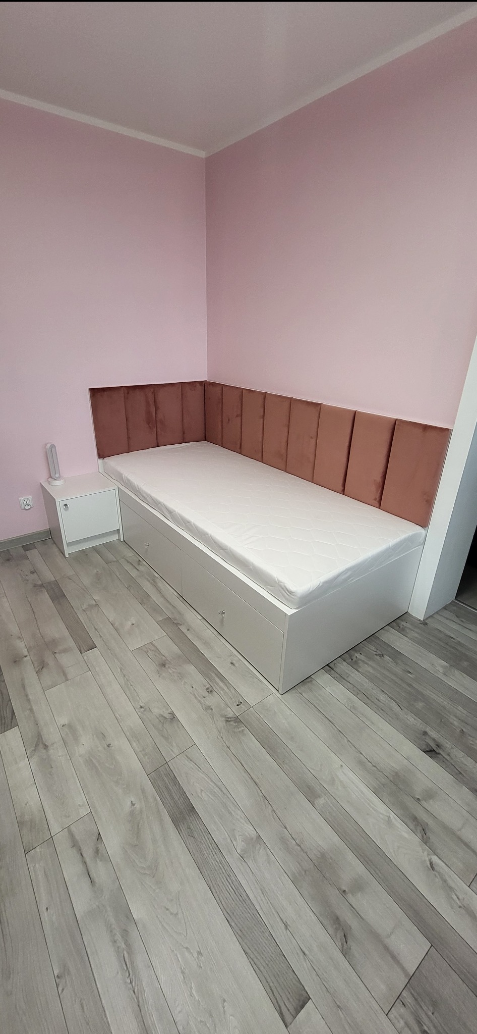 Białe łóżko z szufladami i tapicerowanym, brązowym wezgłowiem na tle różowej ściany. Obok biała szafka nocna. Parkiet w odcieniach szarości.