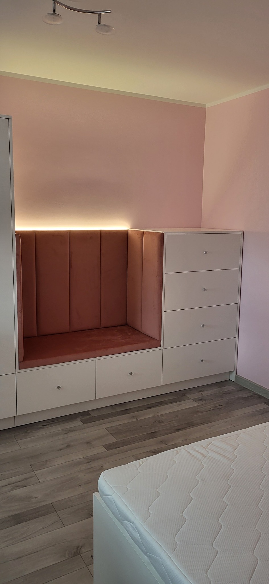 Biała zabudowa meblowa z siedziskiem wykończonym różowym welurem, oświetlona od góry. Szuflady z kryształowymi gałkami, parkiet na podłodze, różowe ściany.