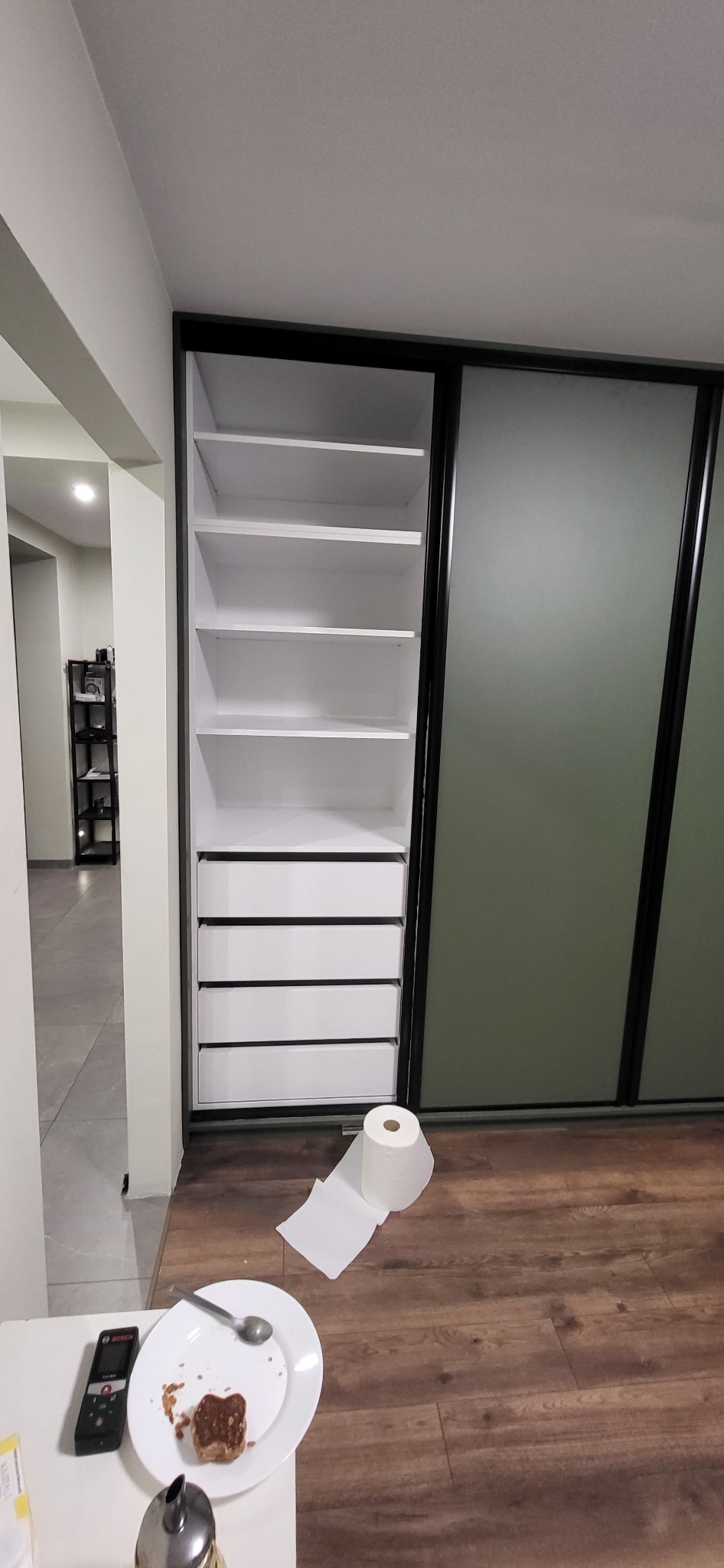 Nowoczesna szafa wnękowa z przesuwnymi drzwiami, białe półki i szuflady, czarne obramowanie. Na podłodze rolka papieru toaletowego i talerz po posiłku.